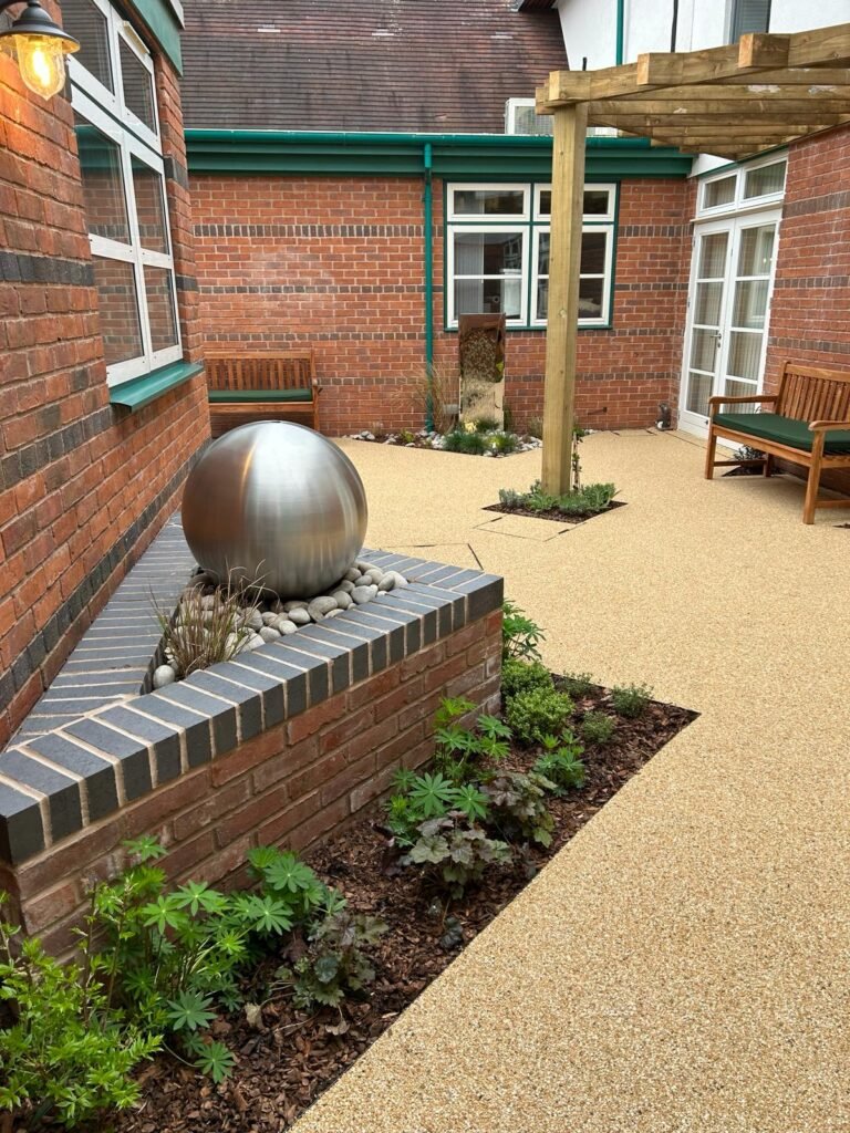 Acorn Hospice in Walsall Commercial Landscaping Project - Resin footpaths, wooden garden structure, outdoor pergola. New planting plan and flower beds. Resin pathways and chrome water feature. Wooden benches. Decorative brick wall. Tree bark planting beds.